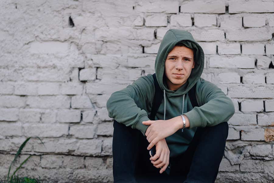 ein junger Mann zw. 16-20 Jahren sitzt vor einer hellen Mauer, er trägt einen Hoodie und hat die Kapuze aufgezogen